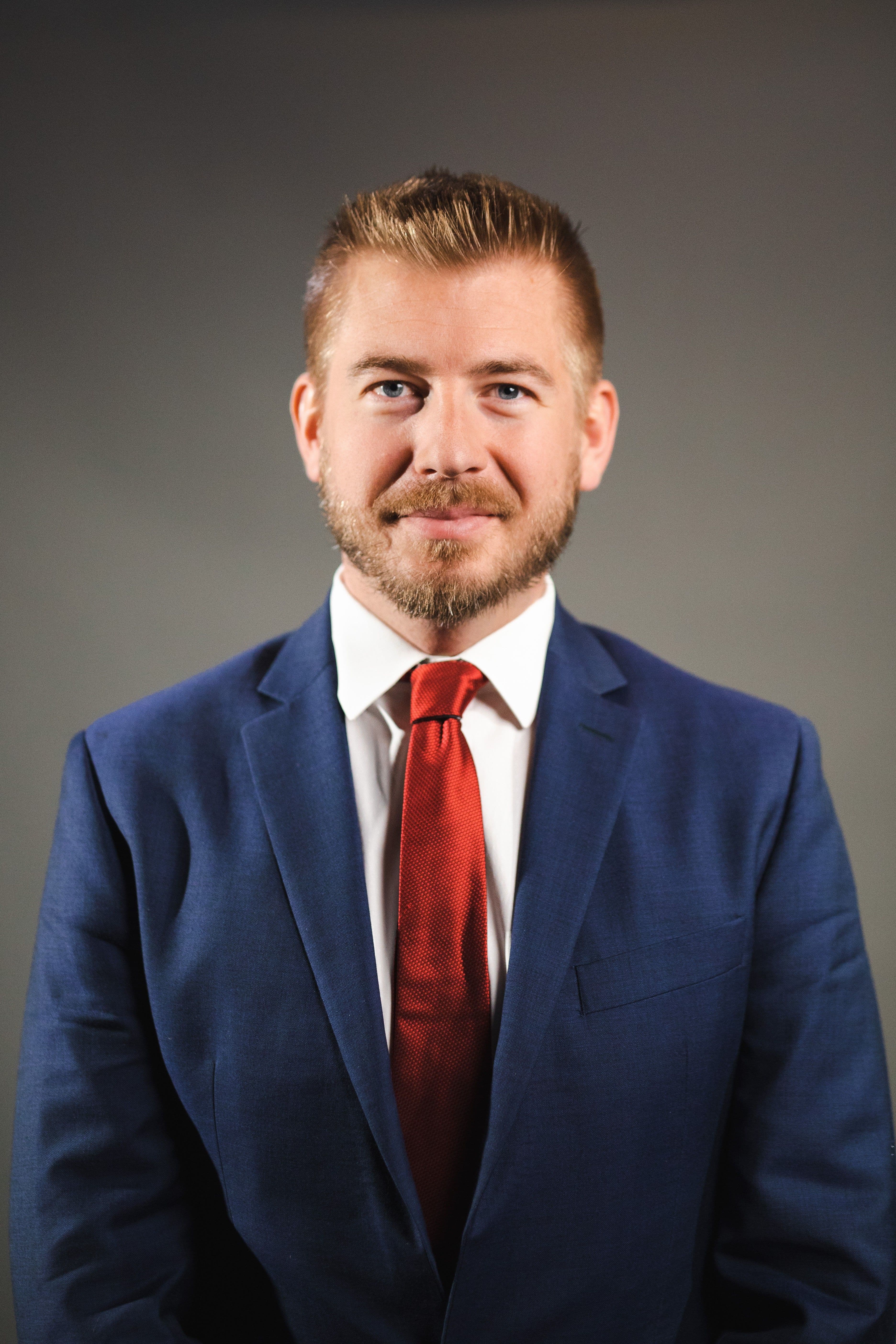 Professional headshot of a man with a beard wearing a blue suit and red tie.