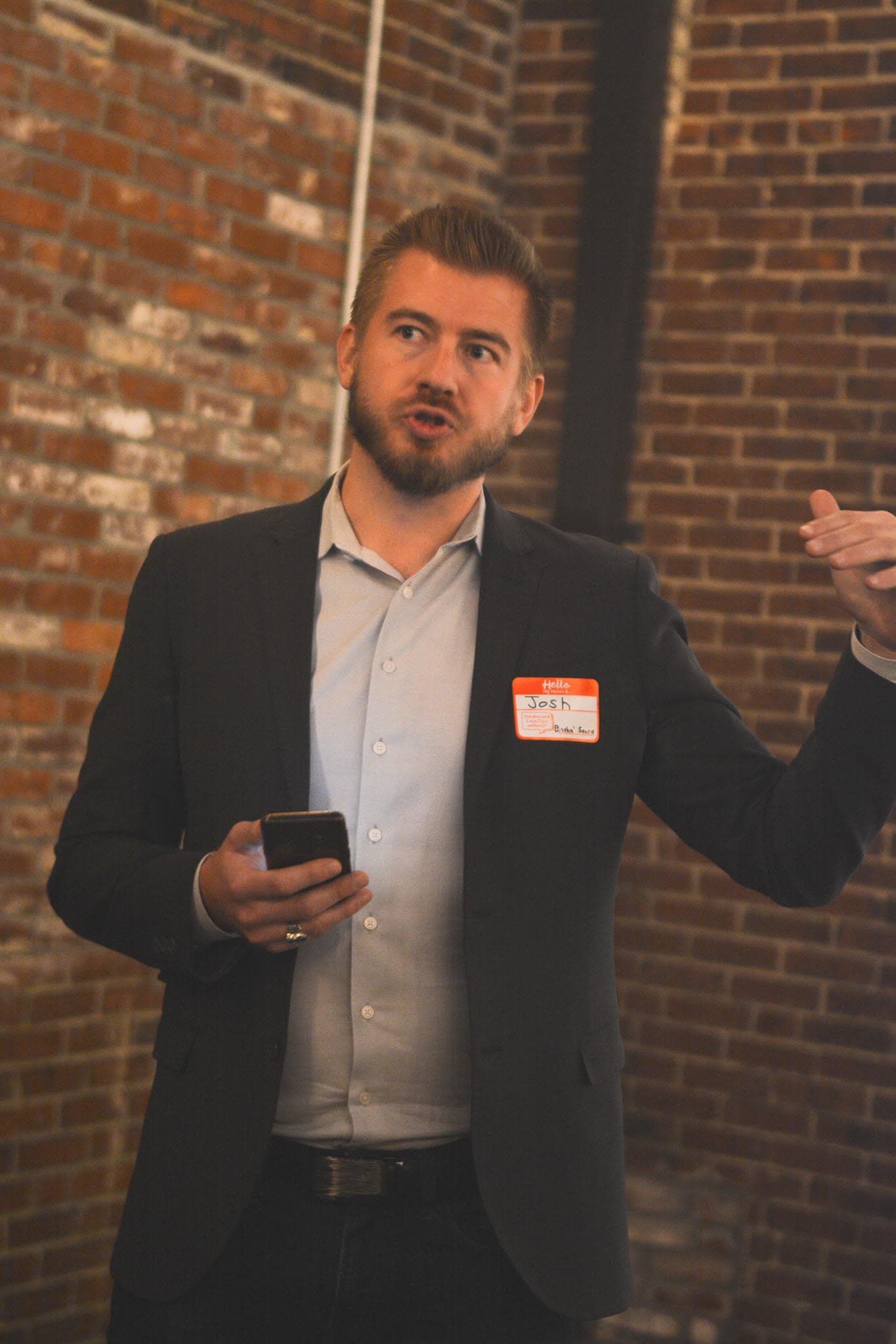 Bearded man named Josh gestures while presenting in a blazer against a brick wall.